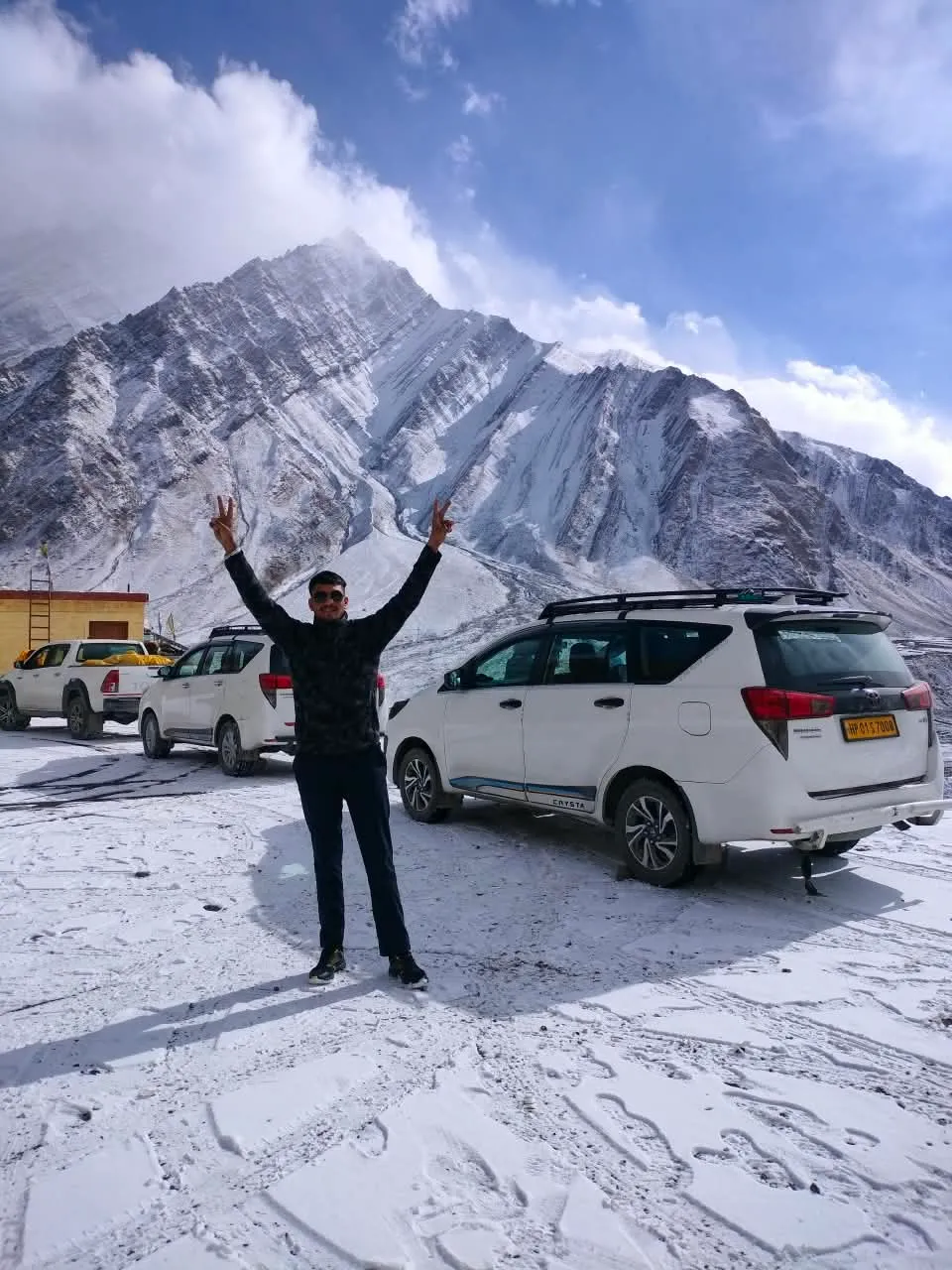 Kulwant with both arms raised in a victory sign on fresh snow, SUVs and a snow-dusted peak behind him